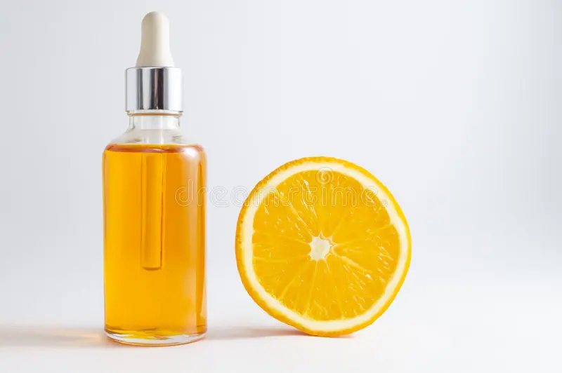 A clear glass dropper bottle filled with orange liquid next to a halved orange on a white background.